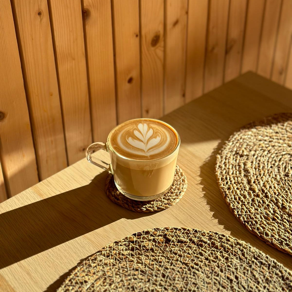 A clear glass cup of latte art coffee sits on a woven coaster on a wooden table, with a warm wood-paneled background and strong sunlight casting shadows.